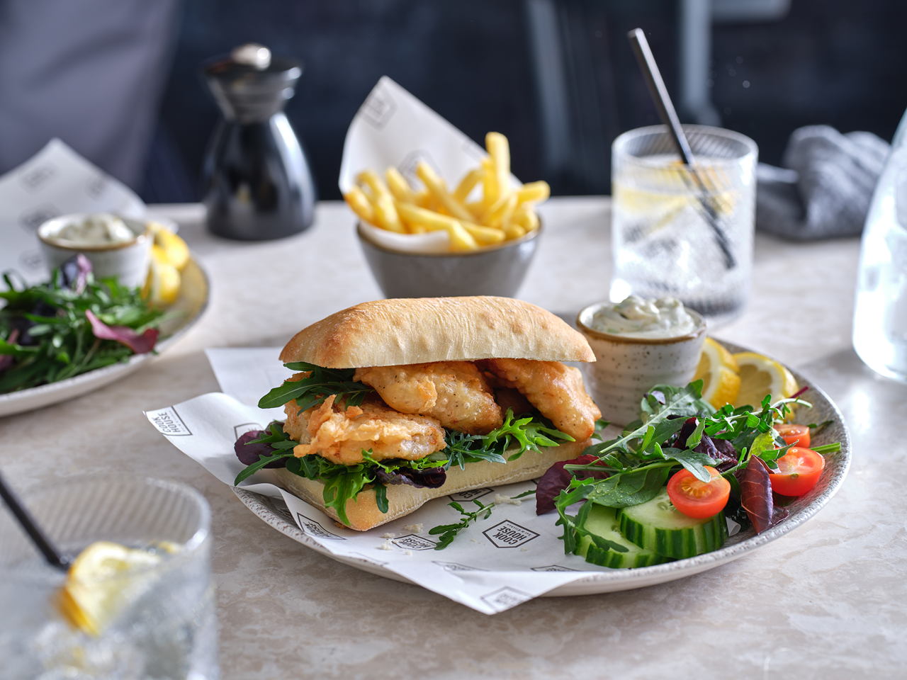 A ciabatta sandwich of battered haddock goujons and green leaves sits on a plate with a side salad, sliced lemon and a pot of tartar sauce. A small bowl of fries can be seen in the background. In the back right and front left there are short glasses containing a clear liquid, lemon slices and a straw.