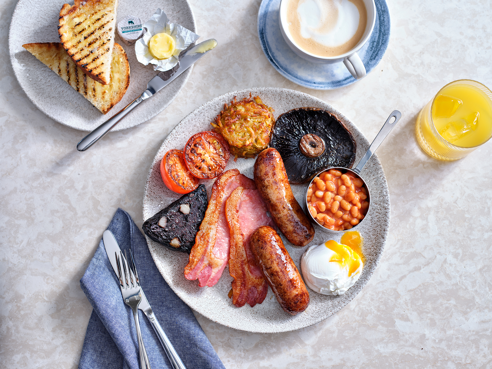 An overhead  photo of a fried breakfast on a plate, including 2 sausages, 2 rashers of bacon, a poached egg, black pudding, tomato, a hash brown and baked beans. Along the top of the frame there's a slice of toast and butter on a plate, a cup of coffee and a glass of orange juice.