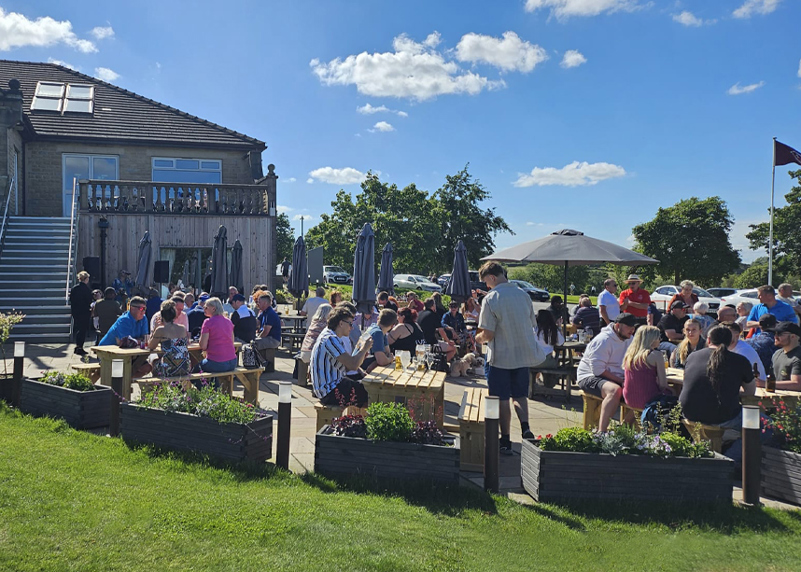 The patio area at Cook House Bar & Kitchen Leeds on a sunny day. The sky is bright blue with a few isolated clouds. The wooden picnic-style tables are occupied by a variety of people. In the foreground there are wooden planters.