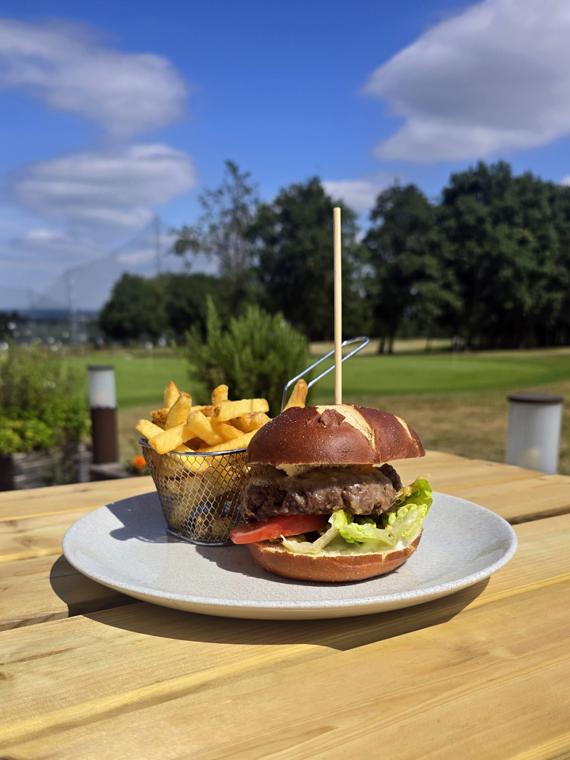 A beef burger in a pretzel bun with lettuce and tomato and a wire basket of fries sitting on a plate placed on an outside wooden table. There are trees in the background and the netting of The Manor Leeds's driving range can be seen.