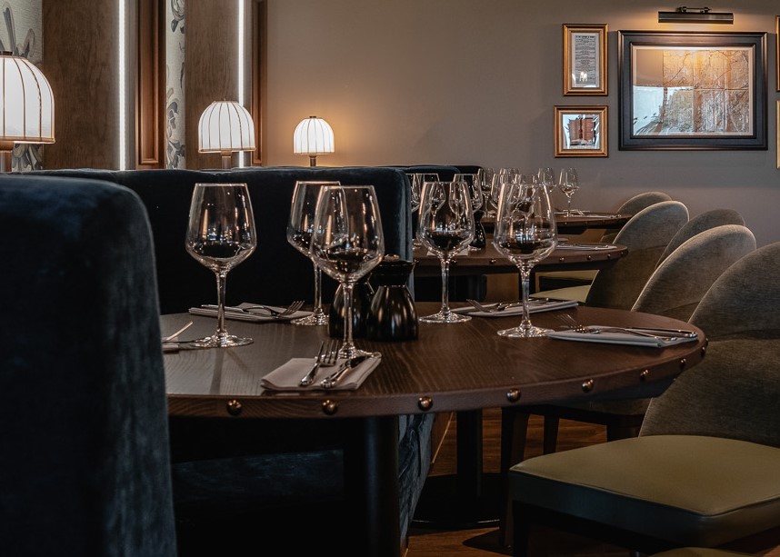 A view of a dining table in Cook House Bar & Kitchen, Leeds. The table is dark wood and round and set for five diners with knives, forks,  napkins and wine glasses. Black salt and pepper mills sit in the centre of the table. The seating on the left hand side of the table is a semi-circular, dark blue velvet banquette and a sage green chair can be seen on the right. There are two more identical table settings in the background.