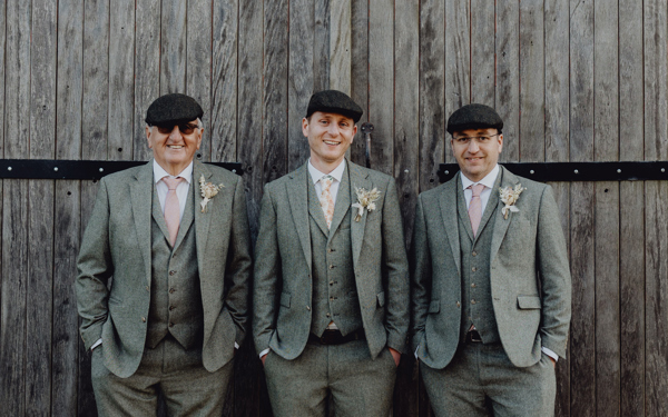Three men dressed smartly in green tweed suits and flat caps stand in front of a wooden barn door, they are smiling.