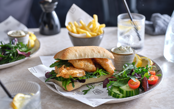 A ciabatta sandwich of battered haddock goujons and green leaves sits on a plate with a side salad, sliced lemon and a pot of tartar sauce. A small bowl of fries can be seen in the background. In the back right and front left there are short glasses containing a clear liquid, lemon slices and a straw.