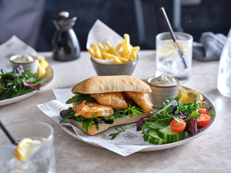 A ciabatta sandwich of battered haddock goujons and green leaves sits on a plate with a side salad, sliced lemon and a pot of tartar sauce. A small bowl of fries can be seen in the background. In the back right and front left there are short glasses containing a clear liquid, lemon slices and a straw.