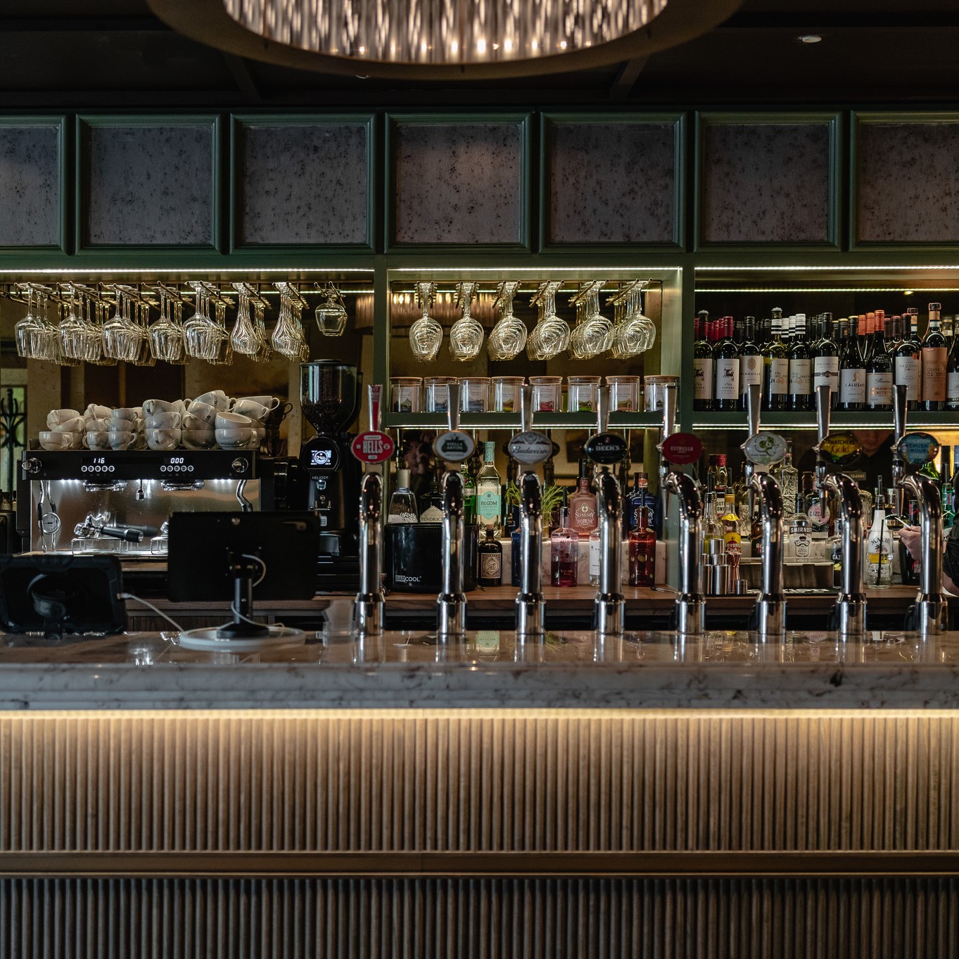 A view of the bar at Cook House Bar & Kitchen. Behind the bar on the left there is a large coffee machine with coffee cups stack on top and wine glasses stored in racks above. The centre shelves hold jars and bottles with more and wine glasses stored in racks above and the shelves on the right hold bottles of wines and spirits. In the foreground, on the marble, there 8 beer pumps. The front of the bar has down lights shining on strips of panelled dark wood.