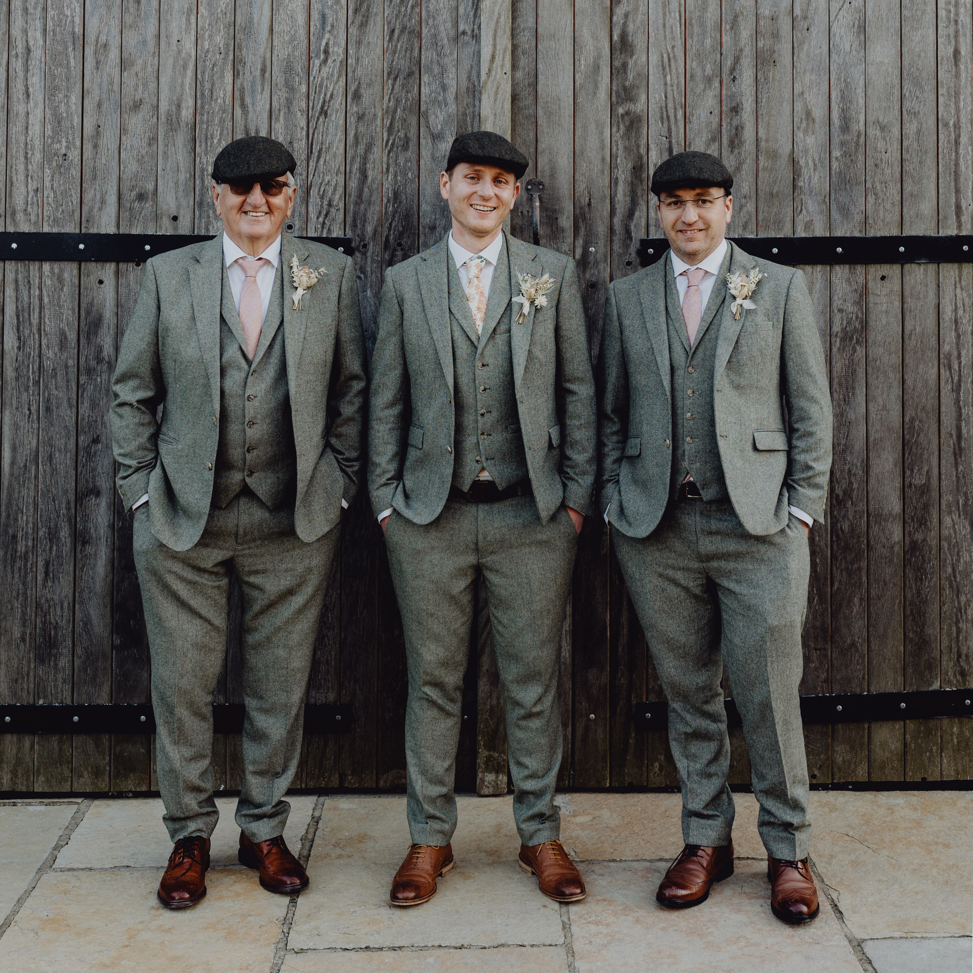 Three men dressed smartly in green tweed suits and flat caps stand in front of a wooden barn door, they are smiling.