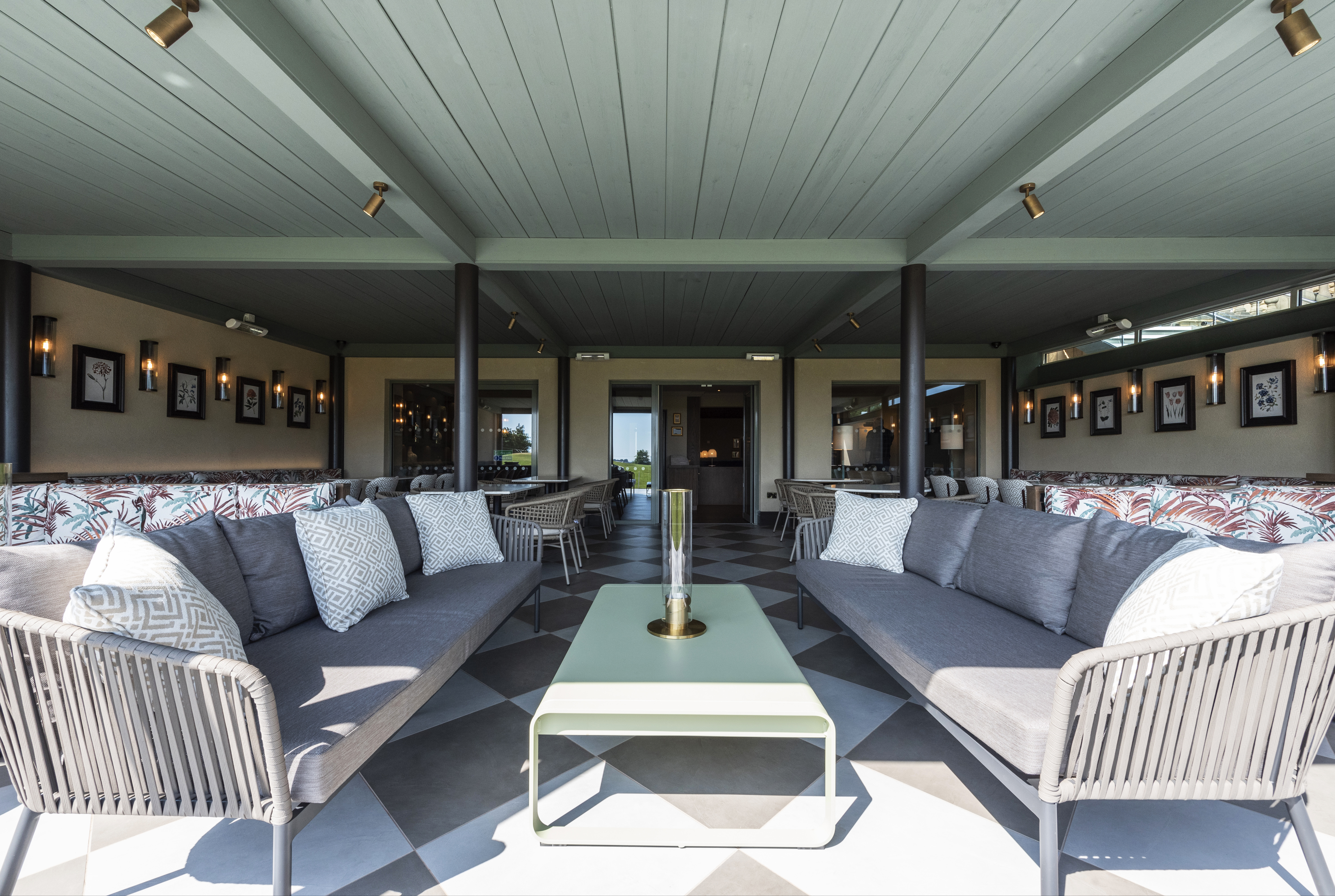 A view into the enclosed terrace area of Cook House Bar & Restaurant, Drighlington. In the foreground there are two sofas facing a low coffee table. Dining tables and chairs can be seen behind and on the rear wall there are glass doors open to the inside bar area. The photo is taken from an open side, the walls to the left and right are solid and the whole space is covered by a wood-panelled ceiling supported by metal pillars.