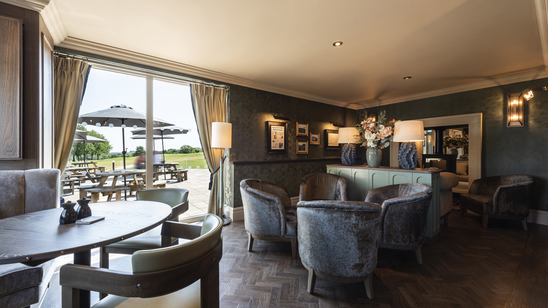 A photo of the snug area of Cook House Bar & Kitchen. A dining table and chairs are on the left of the image and a group of four armchairs are towards the centre in front of a console table holding a large vase of flowers and two lamps. A double sided stove can be seen beyond the table. Picnic benches with parasols can be seen through the large full length windows.