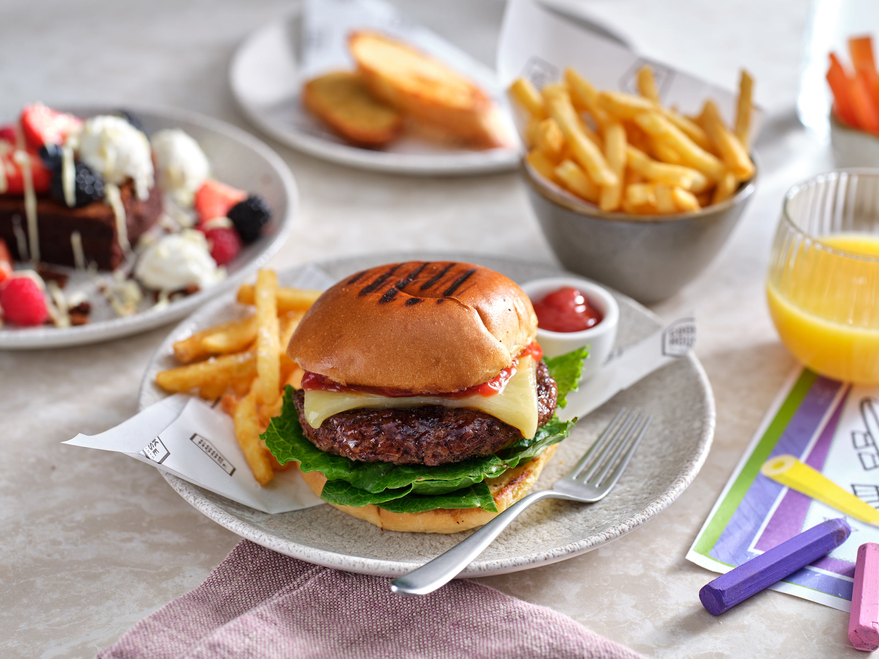 A 4oz beef burger in a bread bun with lettuce and cheese sits on a plate with skinny fries and a small pot of tomato ketchup. A small bowl of fries can be seen in the back right along with a short glass of orange juice and to the back right a chocolate brownie sits on a plate (out of focus).