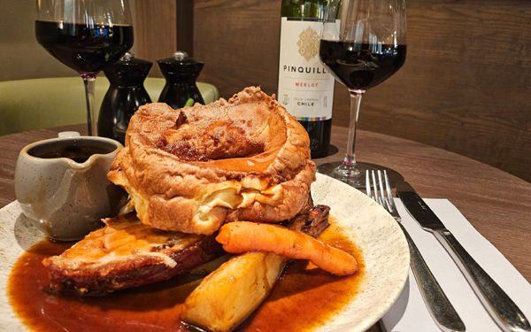 A photo of a roast diner on a plate, including a large Yorkshire pudding, a roasted carrot and parsnip, and a small rug of gravy. A knife and fork lay on a folded napkin to the right hand side and two glasses of red wine can be seen in the background along with the bottle and salt and pepper mills.