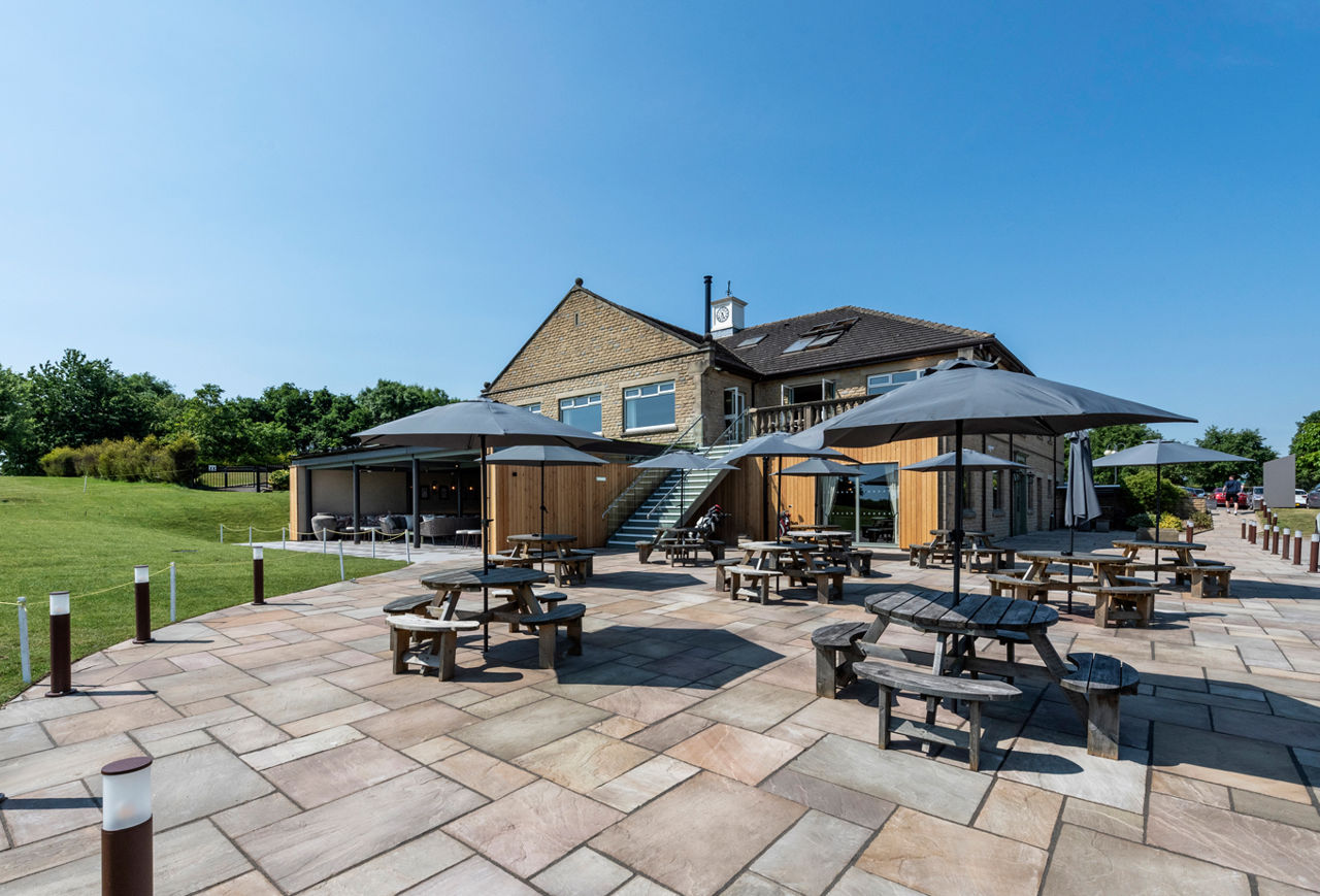 A view of the patio area of Cook House Bar & Kitchen to the rear of The Manor building. In the foreground there is a paved area edged with bollards with nine round picnic-style tables with black parasols in front of a stone building. The exterior of the lower ground floor of the building is wood panelled and there is a metal and glass staircase leading to the upper ground floor. The photo was taken on a sunny day, the sky is bright blue and lawn and trees can be seen to the left.