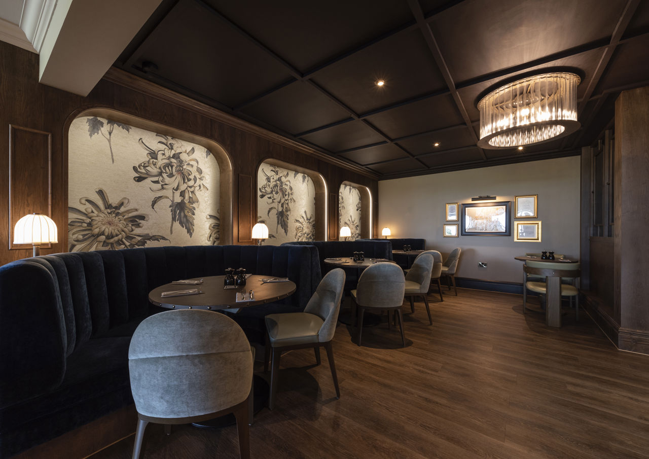 Booth seating at Cook House Bar & Kitchen. Three dining tables can be seen along the wall to the left of the image. Each has semi-circular banquette seating along the wall, and two chairs on the opposite side, each table is round and laid with cutlery and napkins. The floor is dark wood, the ceiling is panelled in dark wood and behind each booth is an alcove featuring light wallpaper with a large floral design. The lighting is low and provided by small lamps between each booth, ceiling spotlights and a large glass light fitting on the right of the image.